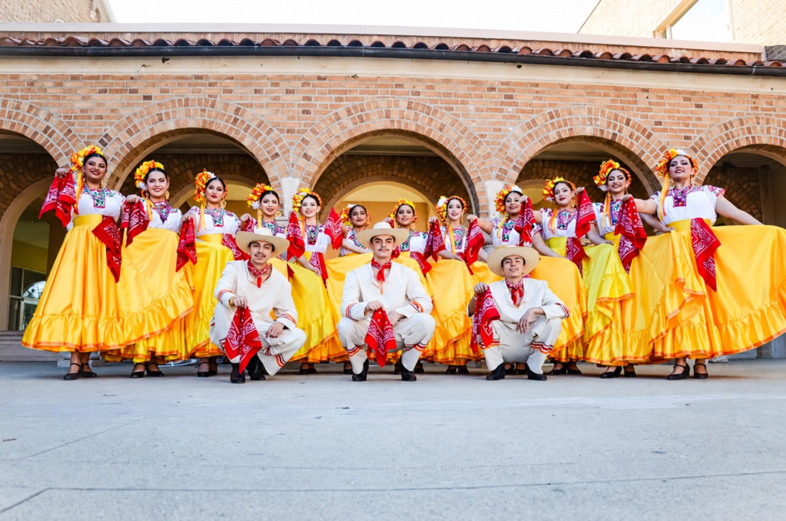 Grupo con vestidos amarillos y trajes blancos frente a arcos de ladrillo